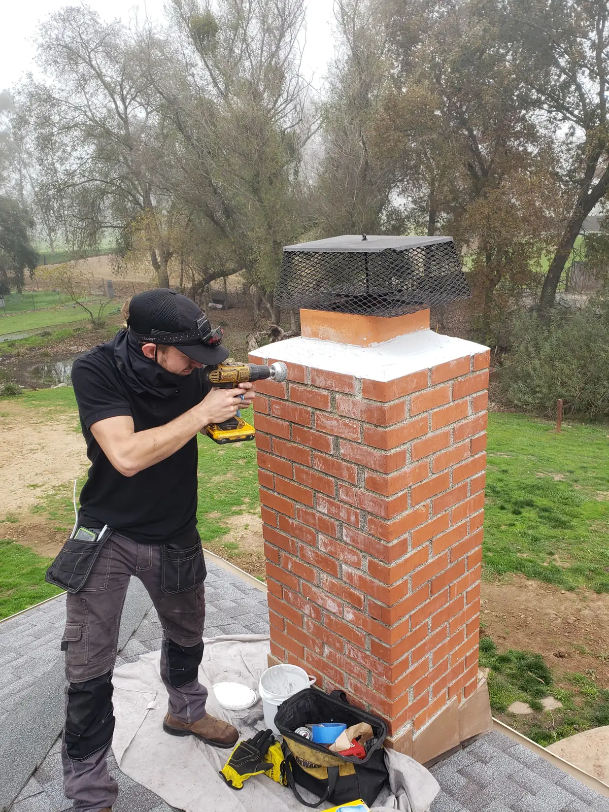 Technician on a roof fitting a stainless rain cap to a rebuilt red brick chimney with a cordless drill