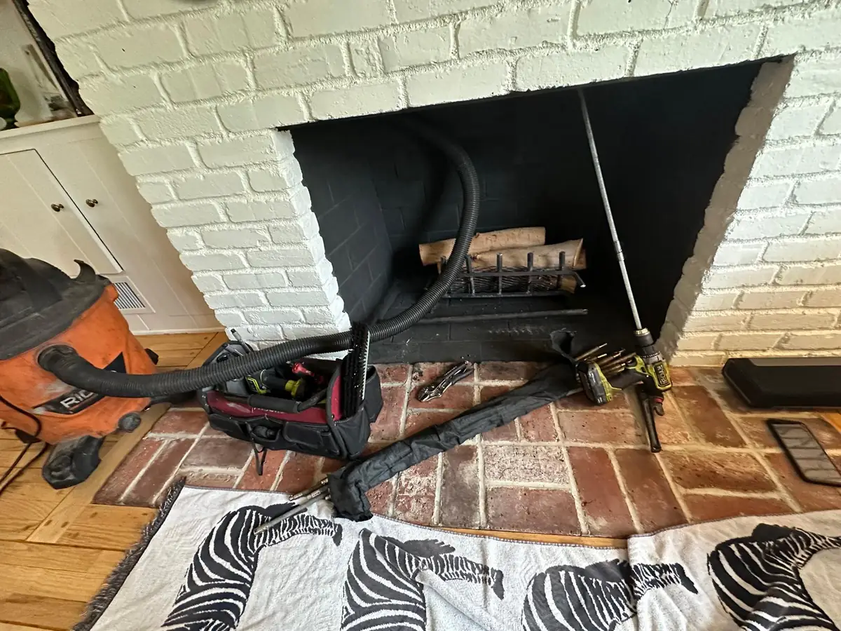 White painted brick fireplace with the damper open and a vacuum hose running into the flue, drop cloth spread across the hearth tiles