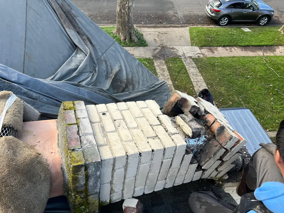Two masons rebuilding the top courses and crown of a brick chimney on a roof, with tarpaulins protecting the work below