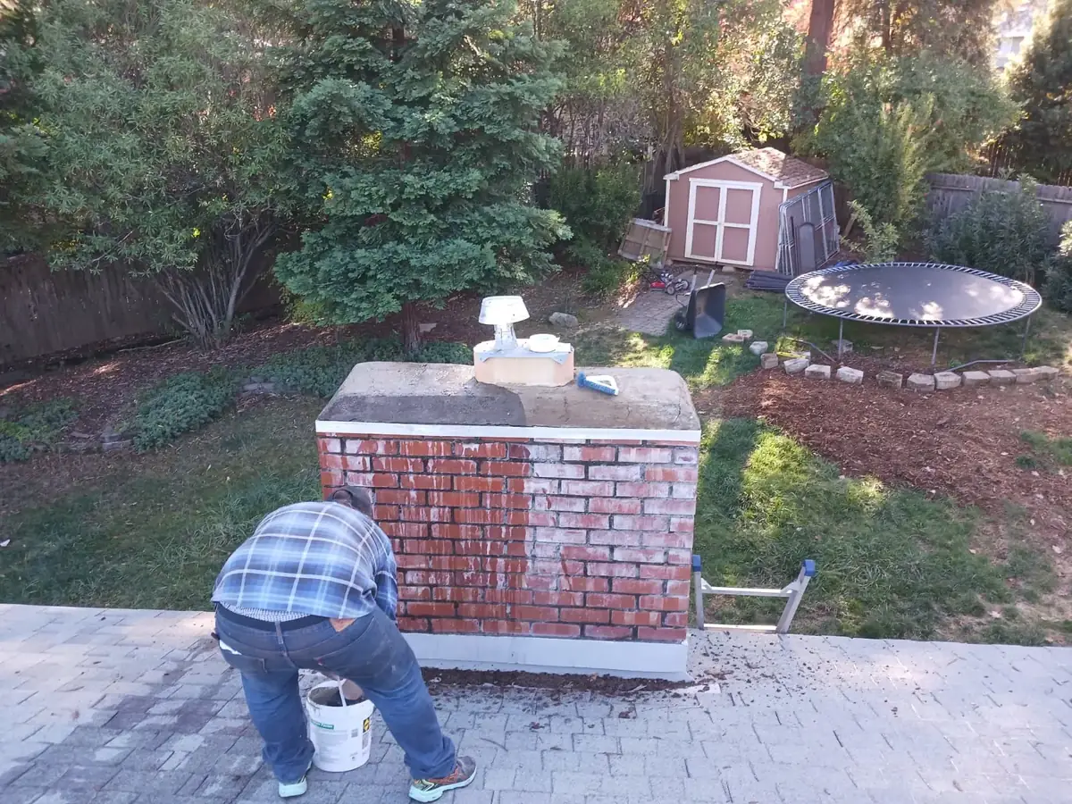 Technician crouched on a shingle roof beside a wide brick chimney with a white cap, inspecting the crown and flashing