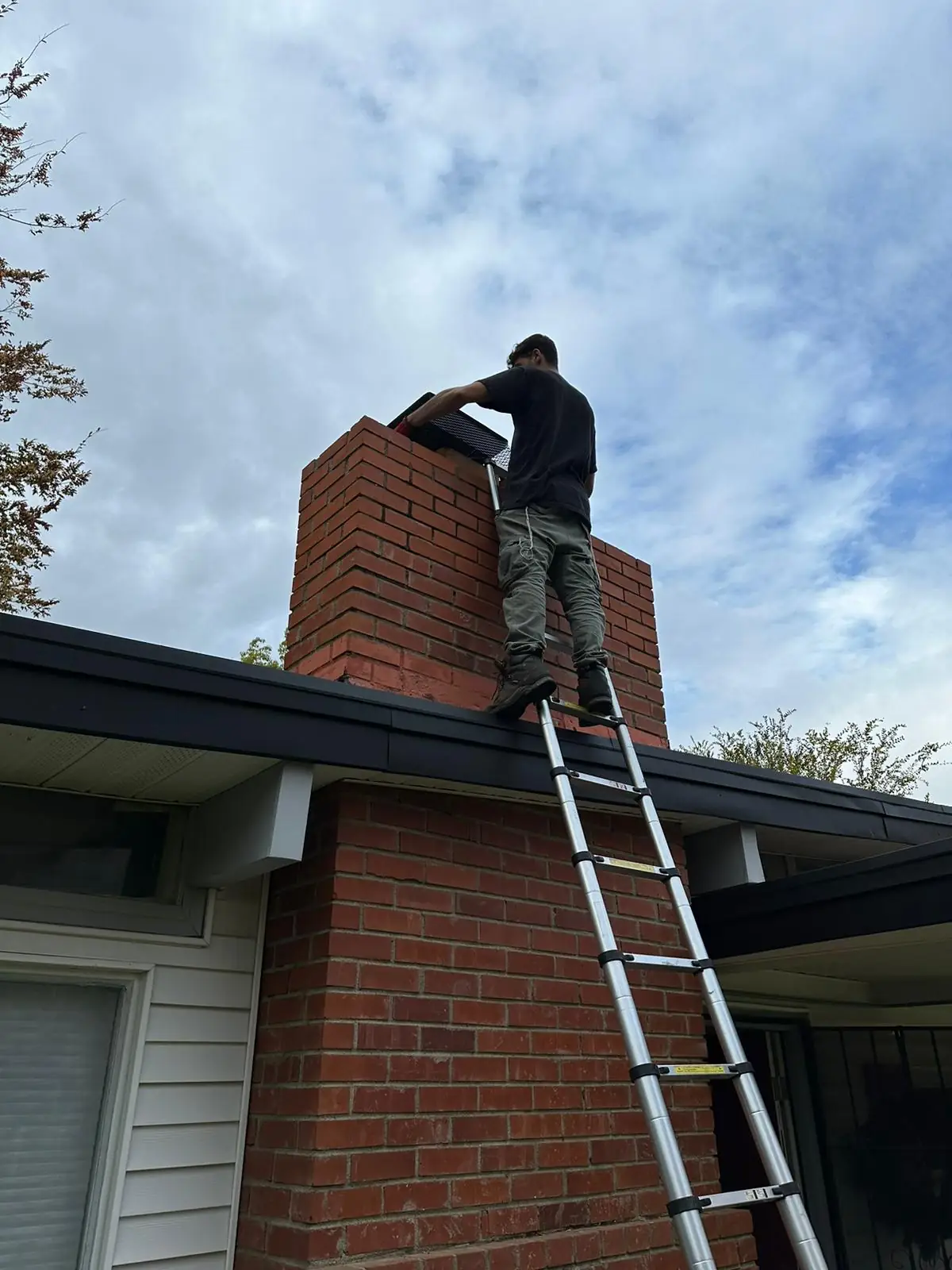 Technician working from an extension ladder at the top of a tall red brick chimney against an overcast sky