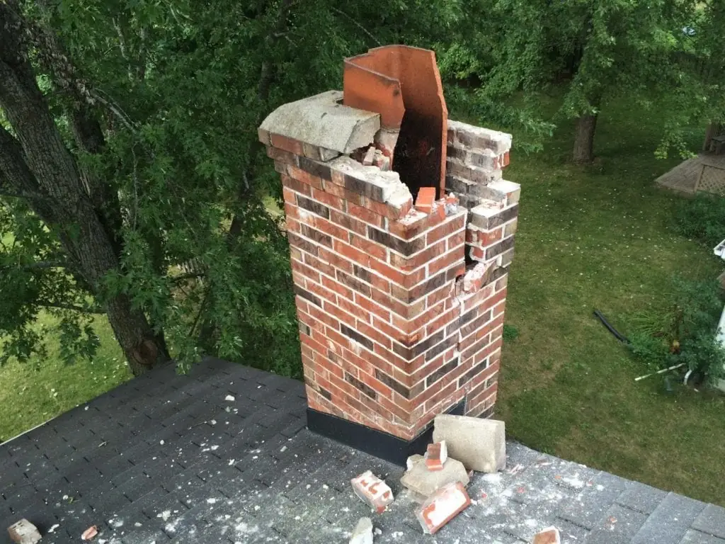 Brick chimney with its crown broken away and loose masonry scattered across the surrounding roof shingles