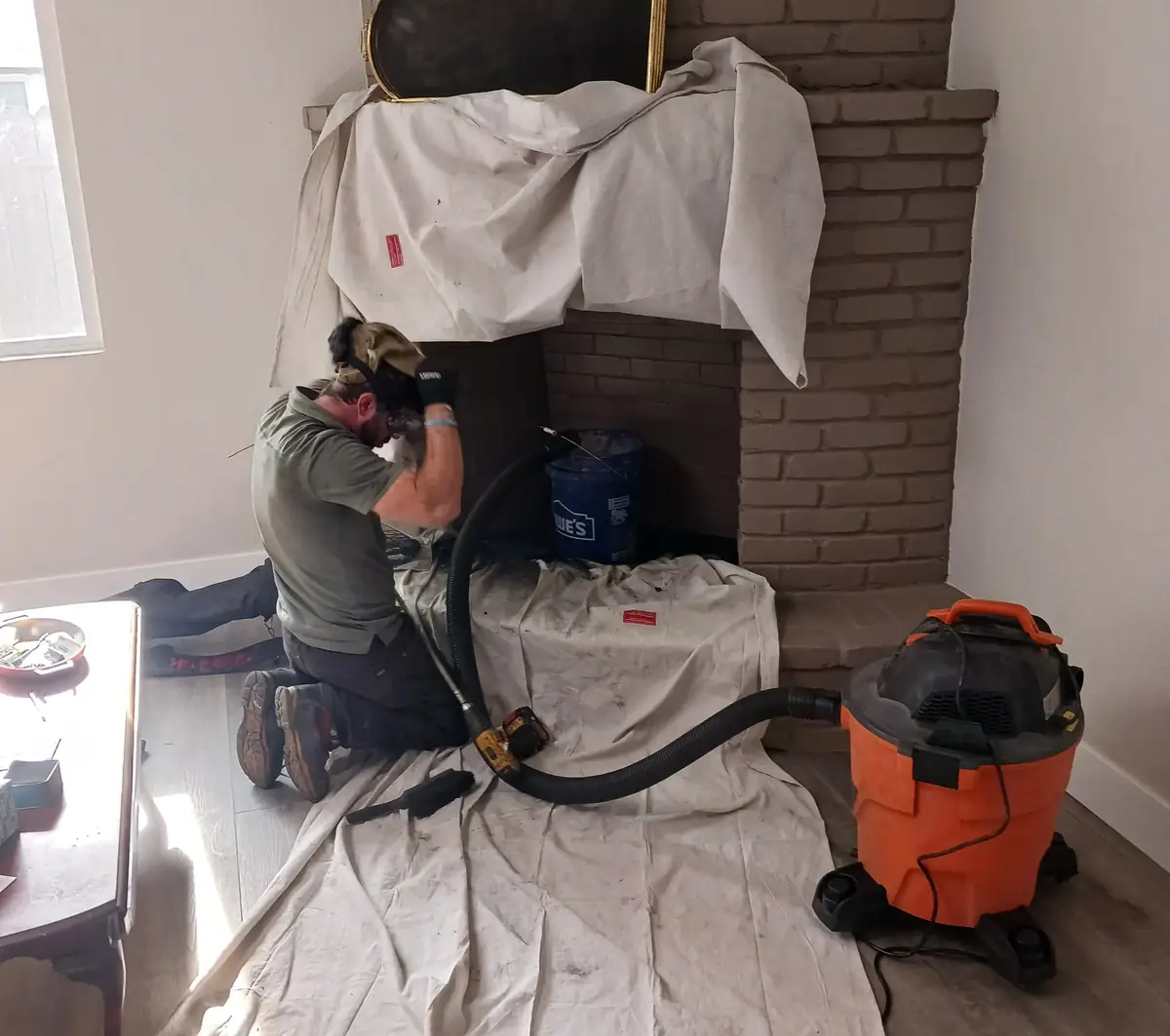 Technician working inside a white-painted brick firebox with dust sheets covering the mantel and floor and a vacuum hose fed into the flue