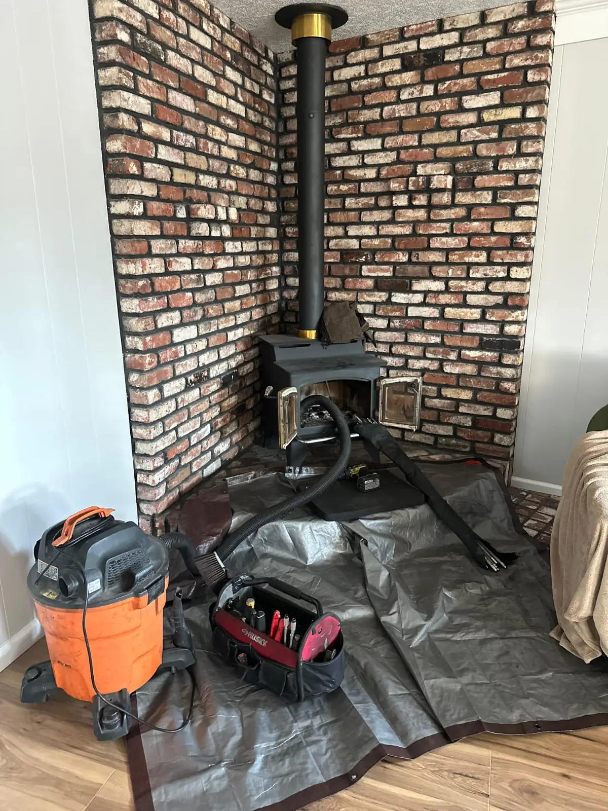 Freestanding wood stove with a black flue pipe rising against an exposed brick wall, protective sheeting and a vacuum on the floor beside it